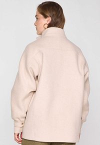 Giacca in misto lana beige con colletto alto, vestibilità comoda e polsini a coste. Presenta una superficie leggermente texturizzata e un design con cuciture minime.