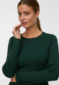Femme aux cheveux châtain clair en queue de cheval portant des boucles d'oreilles en perles et un haut à manches longues texturé vert foncé, touchant son visage.