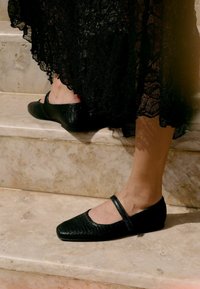 Zapatos Mary Jane planos y negros con cuero texturizado, puntera redondeada y una correa en el tobillo, sobre escaleras de mármol.