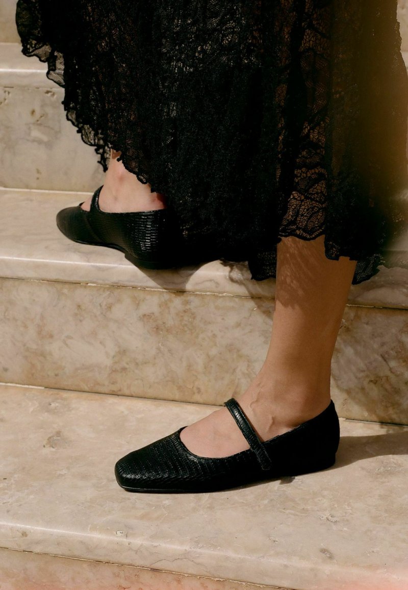 Zapatos Mary Jane planos y negros con cuero texturizado, puntera redondeada y una correa en el tobillo, sobre escaleras de mármol.