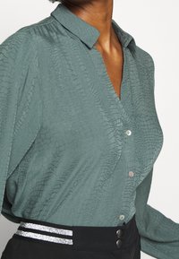 Chemise verte à boutons avec un motif texturé en écailles, dotée d'un col et de boutons nacrés. Associée à un pantalon noir avec des rayures argentées.