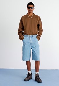 Brun genser, lyseblå denimshorts, svarte tursko og solbriller. Genseren har rund hals og en avslappet passform.