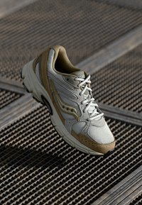 Lys grå og beige treningssko med mesh-overdel, polstret krage, lisser og gummisåle. Har subtile teksturmønstre og merkevare.