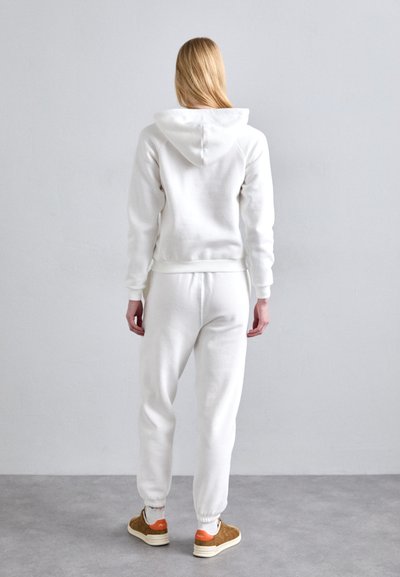 Vit huvtröja och matchande joggers i mjukt material. Har ribbade muddar och midja. Bruna skor med orange detaljer.