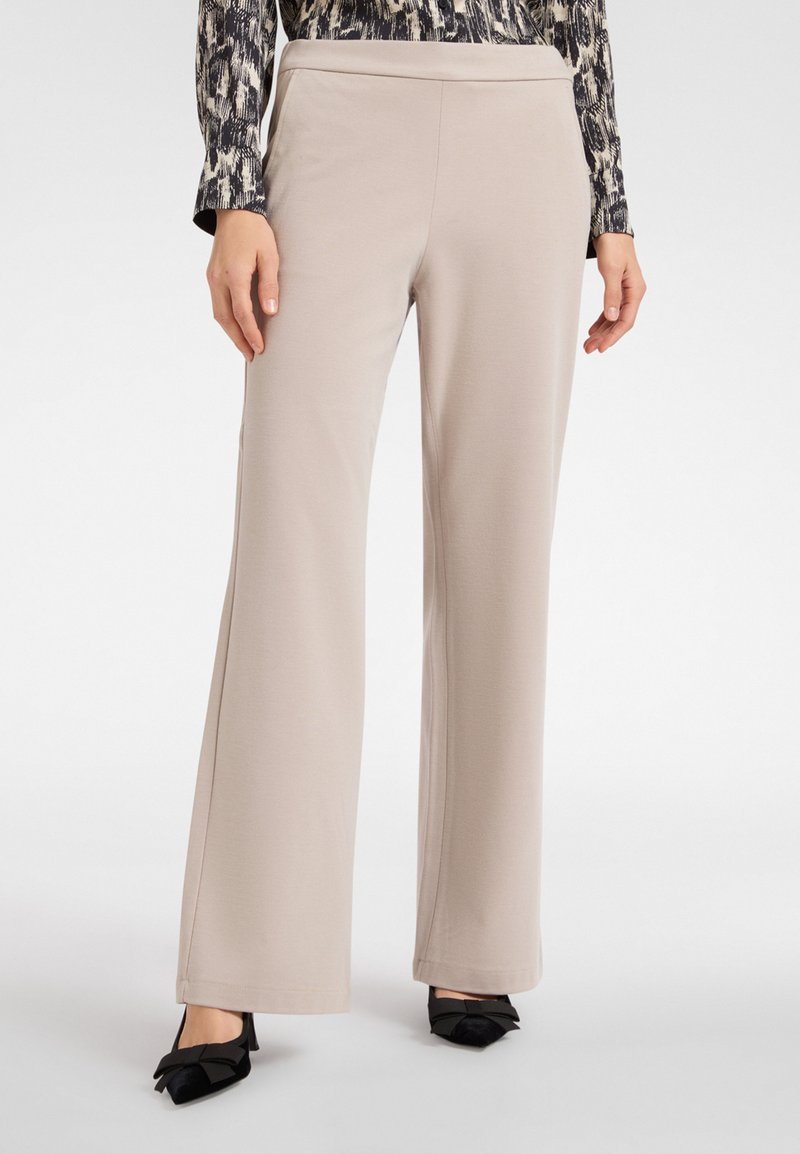 Beige wijde broek met een zachte textuur, uitgerust met een hoge taille. In combinatie met zwarte puntige schoenen versierd met strikken.