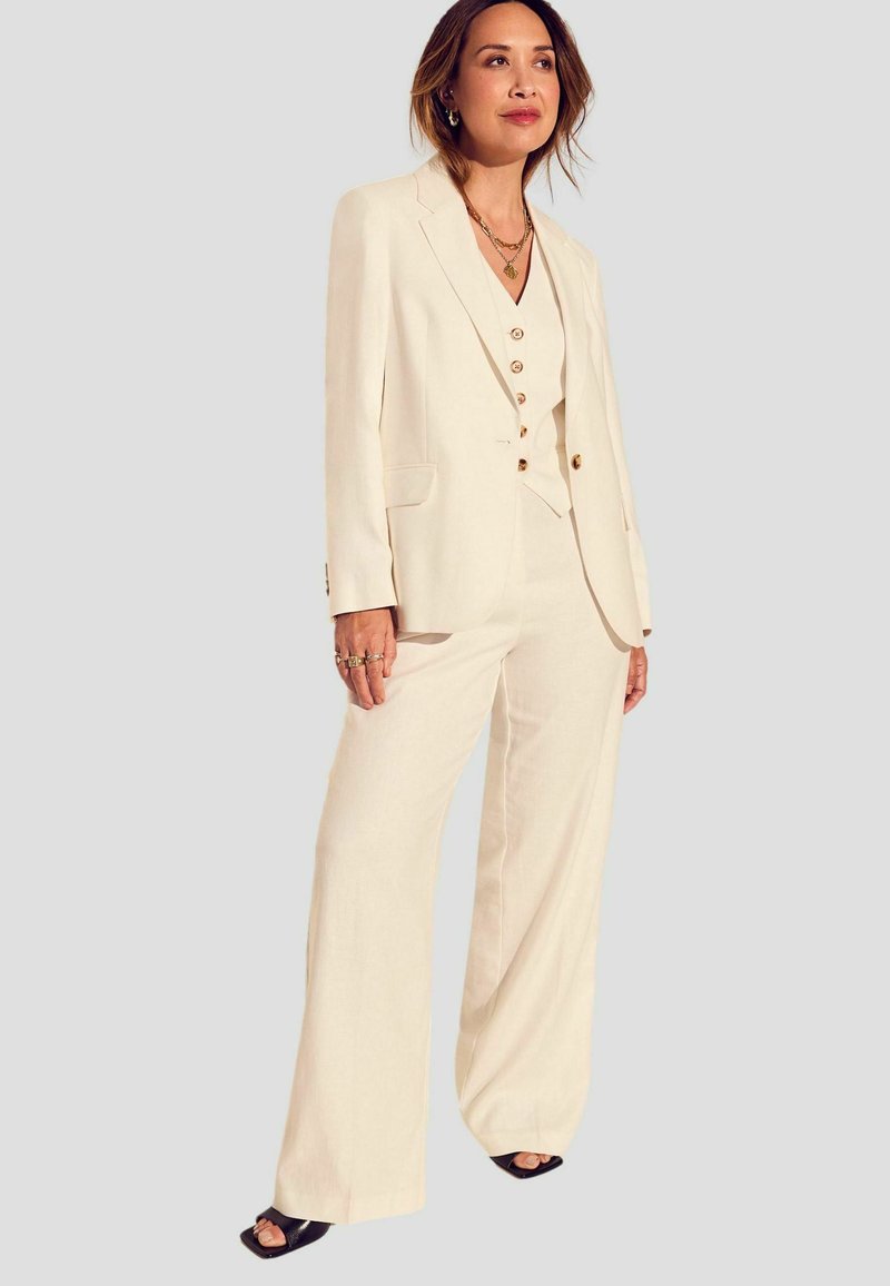 Costume sur mesure couleur crème avec un long blazer et un pantalon à jambe large, tissu texturé, et boutons contrastants sur le gilet. Chaussures noires à bout ouvert.