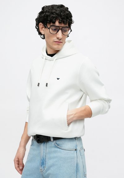 Balta hoodie su kanguros tipo kišene, raišteliu po gaubtu ir mažu logotipu ant krūtinės. Derinama su šviesiai mėlynais džinsais ir juodu diržu.