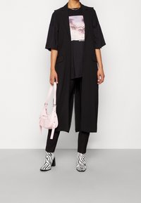 Černá dlouhá vesta přes černé tričko s grafickým motivem, černé kalhoty, růžová kabelka a kotníkové boty se zebra potiskem. Hladké textury a střihaný design.