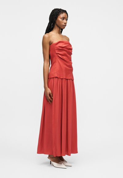 Nő hosszú fonatokkal, pánt nélküli, fodros piros felsőben és hozzá illő hosszú szoknyában, fehér, hegyes orrú, magassarkú cipővel, oldalra fordulva áll.