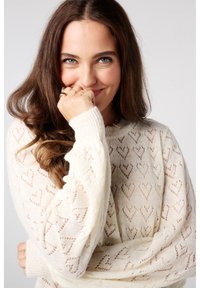 Pull en tricot crème avec des motifs en forme de cœur, design en dentelle texturée, et poignets ajustés. Le mannequin pose avec une expression espiègle, mains près du visage.