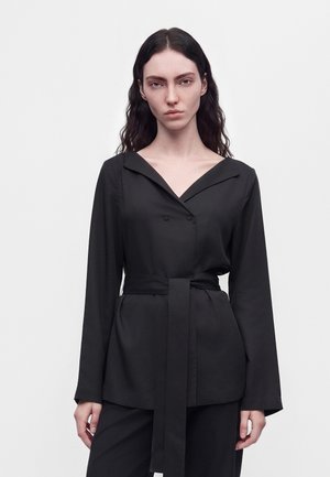 Un mannequin aux longs cheveux foncés porte un blazer noir ceinturé et un pantalon, devant un fond clair uni.
