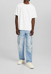 Vit bomullst-shirt med korta ärmar, kombinerad med ljusblå cargojeans med flera sidofickor och vita sneakers.