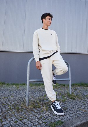 Cremefarbener Strickpullover mit schwarzen Akzenten, kombiniert mit passenden cremefarbenen Jogginghosen. Schwarz-weiße Sneaker runden das Outfit ab.