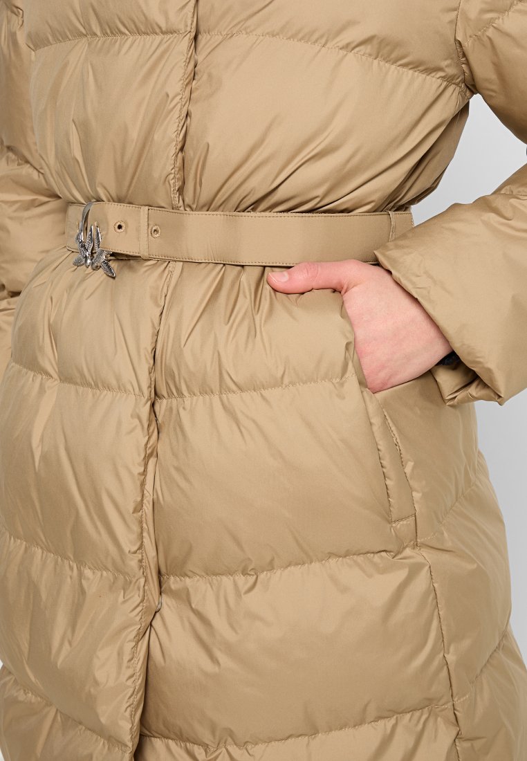 Cappotto imbottito beige con cintura dotata di fibbia argentata, mano inserita nella tasca laterale, primo piano sulla parte centrale e sulla manica.
