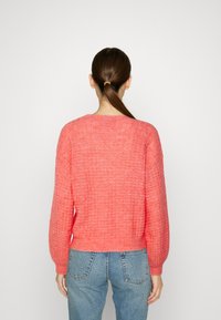 Korallfarbener Strickpullover mit strukturiertem Muster, Rundhalsausschnitt und langen Ärmeln; kombiniert mit hochgeschnittenen hellblauen Jeans.
