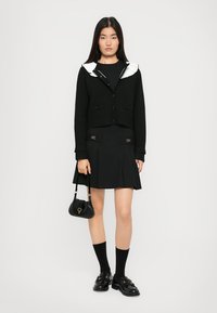 Cardigan nero corto con colletto bianco, abbinato a una gonna nera con dettagli dorati, calze nere fino al ginocchio e mocassini neri. Comprende una borsa piccola.