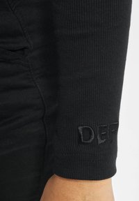 Crna majica dugih rukava s rebrastom teksturom i prilagođenim dizajnom. "DEF" izvezeno crnim koncem na manžetama dodaje suptilni detalj brendiranja.