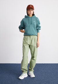 Teal katoenen hoodie met een voordtas, gecombineerd met lichtgroene baggy cargo shorts en witte sneakers. Bruine pet met geborduurd detail.