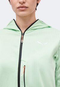 Κοντινό πλάνο προσώπου που φοράει ανοιχτό πράσινο μπουφάν με κουκούλα, μαύρο φερμουάρ και μικρό λευκό λογότυπο στο στήθος.