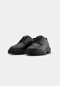 Pantofi din piele neagră cu vârf rotunjit, talpă robustă cu striații și design cu șireturi. Au o textură netedă și brandizare minimă pe lateral.