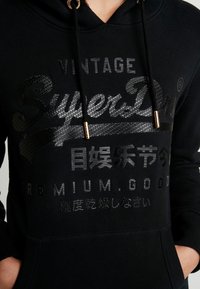 Felpa nera con hardware dorato, caratterizzata da testo in rilievo nero, tra cui "Vintage SuperDry" e vari caratteri, realizzata in cotone e con polsini a costine.