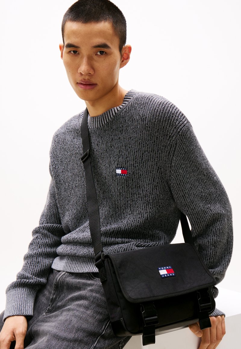 Maglione grigio a coste con scollatura rotonda. Borsa a tracolla nera in tessuto con patch del logo e cinturini con fibbia. Stile casual e confortevole.