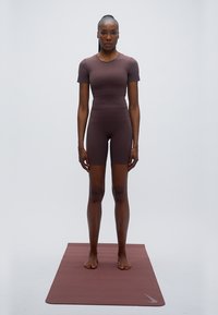 Καφέ αθλητική στολή από ελαστικό υλικό· μπλούζα με κοντά μανίκια και σορτς μέχρι το μέσο μηρού. Το μοντέλο στέκεται ξυπόλυτο σε ένα ταίριαστο χαλάκι γιόγκα.