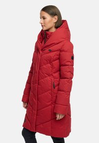 Rød polstret jakke med diagonal quiltet mønster, stor hætte og lynlåse på sidelommerne. Har dekorative knapdetaljer på ærmerne.