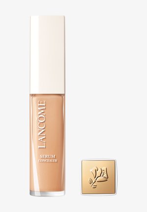 Lancôme serum concealer in a tall, clear tube with a beige liquid and a white cap; accompanied by a small square gold compact with floral design.