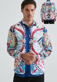 Chemise à boutons multicolore avec des motifs complexes d'oiseaux et de feuillage. Fabriquée en tissu léger avec un panneau avant bleu contrastant.