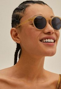 Occhiali da sole rotondi con una montatura gialla traslucida e lenti scure. La modella ha i capelli intrecciati e indossa un top marrone.