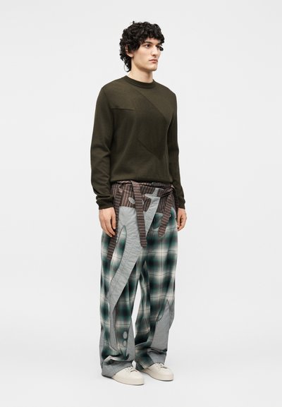 Άνδρας με σγουρά μαλλιά που φοράει ελιά πουλόβερ και φαρδιά, πολυτεμαχισμένα καρό παντελόνια με λευκά αθλητικά, στέκεται μπροστά σε απλό φόντο.