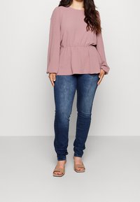 Femme portant un blouse mauve à manches longues avec taille cintrée, un jean skinny bleu foncé et des sandales à talons ouverts beiges, se tenant sur un fond uni.