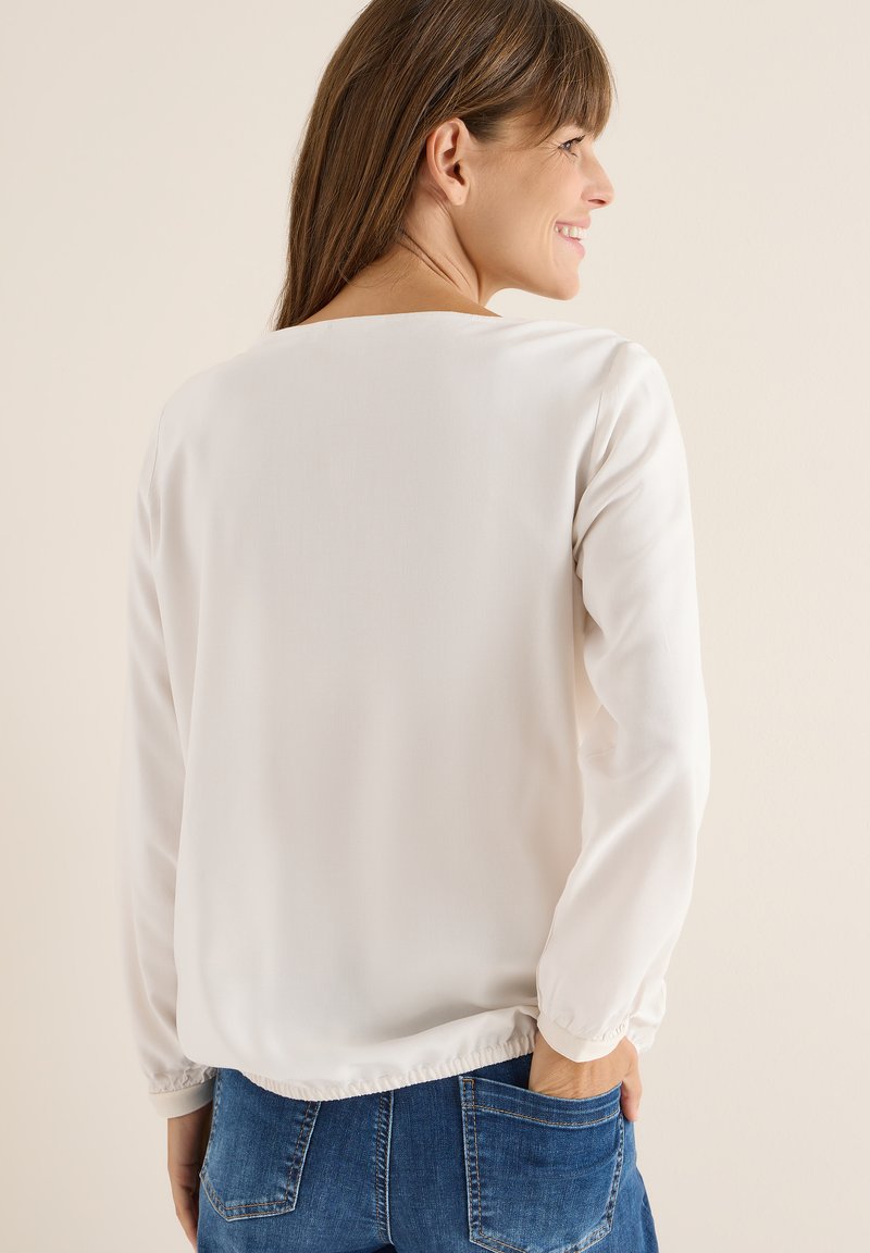 Hvid langærmet bluse med en rund halsudskæring og elastisk kant, lavet af letvægtsstof, parret med blå denim jeans.