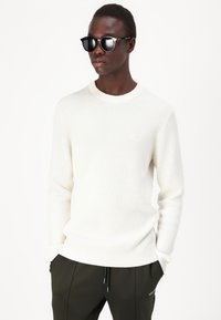 Maglione a coste bianche con scollatura a girocollo, indossato con pantaloni scuri. Il modello indossa anche occhiali da sole neri.
