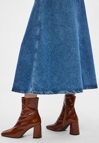 Brūnas lakotas ādas potīšu zābaki ar kvadrātveida purngalu un plata papēdi, valkāti kopā ar džinsa svārkiem, kas rāda taisnu apakšmalu.