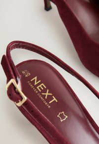 Burgundy mockasins med hög klack och en tunn ankelrem, guld "NEXT FOREVER COMFORT" branding och spetsig tån design.