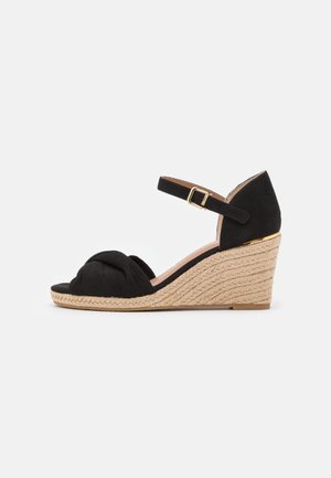 Sort kile sandal med fletet jutesål, knyttet frontstropp og ankelrem med guldlås, på hvid baggrund.