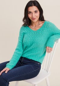 Türkiser gestrickter Pullover mit einem strukturierten Muster, V-Ausschnitt und langen Ärmeln, kombiniert mit dunkelblauen Jeans. Das Modell sitzt auf einem weißen Stuhl.