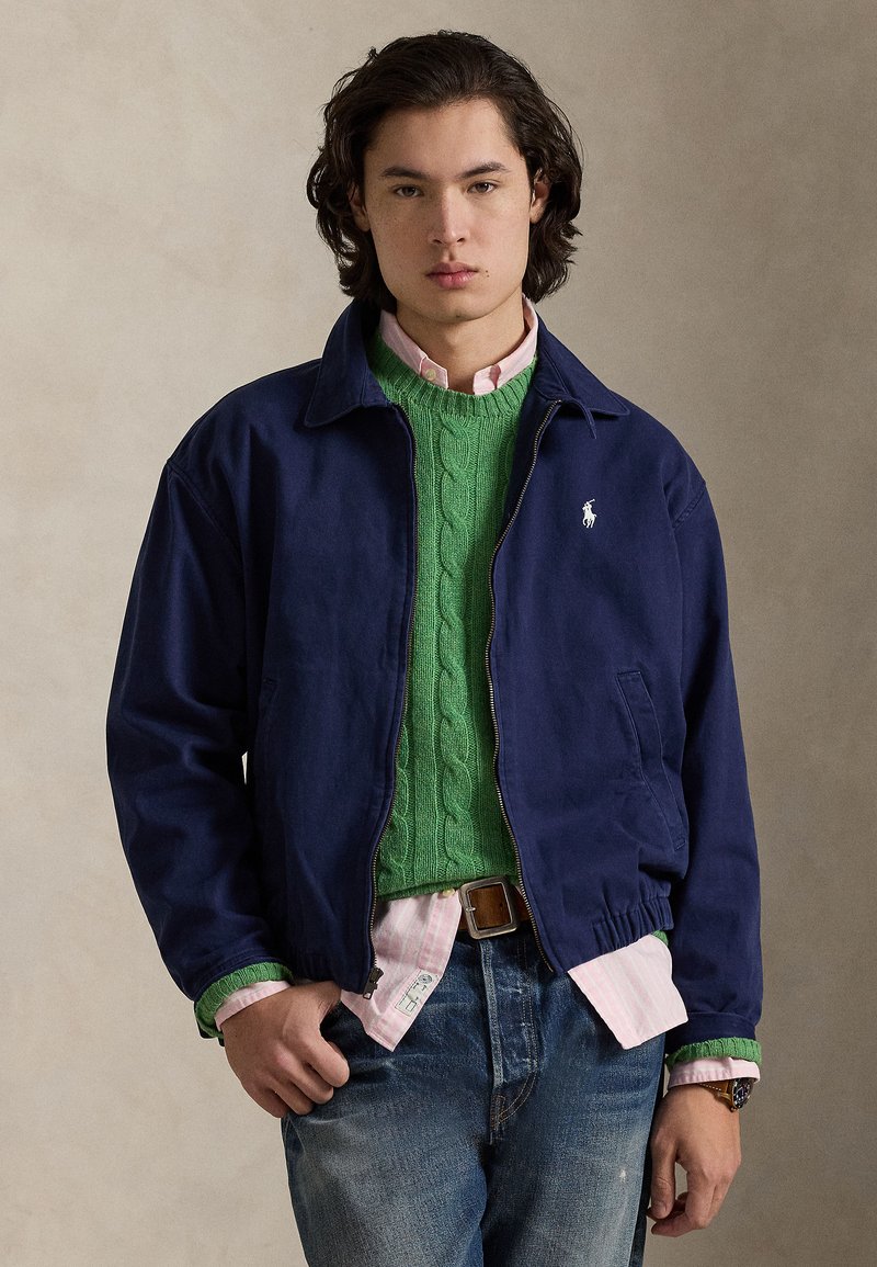 Ung mand iført en navy jakke over en grøn kabelstrikket sweater og lyserød skjorte med blå jeans, stående mod en ensfarvet beige baggrund.