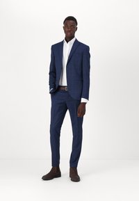 Navyblau karierter Anzug mit taillierter Passform, ausgestattet mit einem Reverskragen, einem weißen Dress-Shirt und braunen Stiefeletten. Glatte Stofftextur.