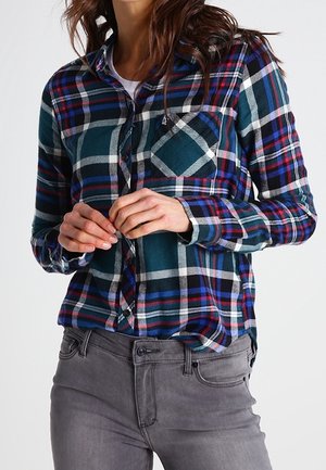 Kvinde iført en blå, grøn og hvid ternet skjorte med knapper over en hvid top og grå jeans, hænderne foldet nær taljen.