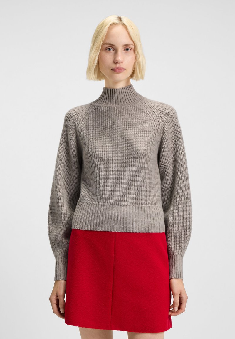 Blonde woman wearing a gray ribbed turtleneck sweater and a bright red skirt standing against a plain white background.