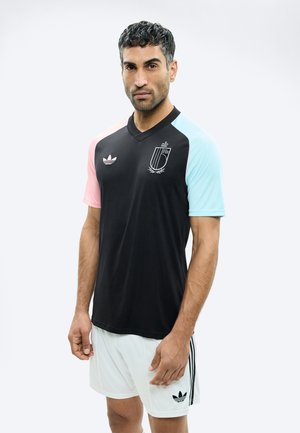 Homme portant un maillot de sport noir avec des manches roses et bleu clair, ainsi qu'un short blanc, debout devant un fond clair.