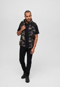 Fleeceväst med ett kamouflagemönster i mörkgröna, svarta och blå toner. Har dragkedja och hög krage. Kombineras med svarta jeans och stövlar.