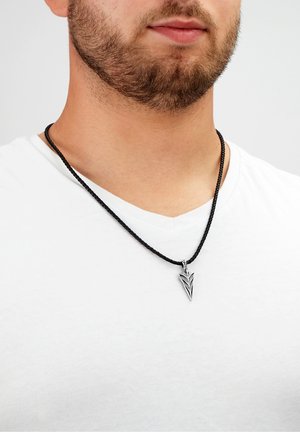 Schwarz geflochtene Halskette mit einem silbernen Pfeilspitzenanhänger, der geometrische Details aufweist, getragen über einem weißen Hemd.