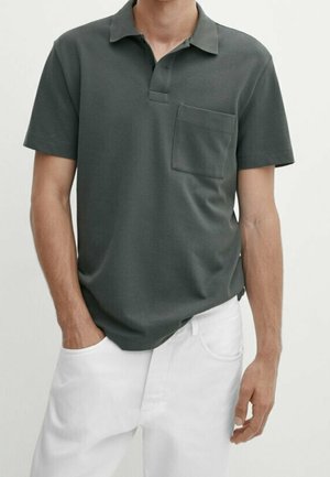 Mężczyzna ubrany w ciemnoszary polo z krótkim rękawem i kieszenią na piersi oraz białe spodnie, stojący z jedną ręką w kieszeni.