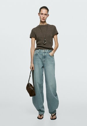 Donna in piedi che indossa un top marrone lavorato a maglia a maniche corte, jeans larghi celeste chiaro, sandali neri, una lunga collana con pendente e tiene una borsa marrone.