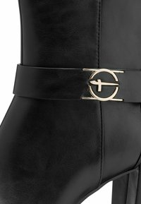Bottine en cuir noir avec une large sangle ornée d'une boucle dorée circulaire et une texture mate lisse, mettant en valeur le design élégant de la bottine.