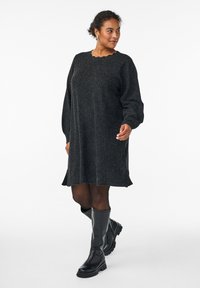 Robe noire tricotée avec un décolleté festonné et de longues manches bouffantes, présentant une coupe décontractée et un ourlet effiloché. Associée à des bottes noires jusqu'aux genoux.